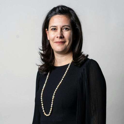 A woman with long dark hair wearing a black dress and a long pearl necklace stands against a plain light gray background, looking at the camera with a slight smile.