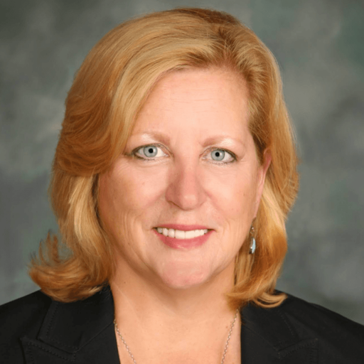 A woman with shoulder-length blonde hair, light eyes, and a friendly expression, wearing a dark blazer and a light necklace, poses in front of a gray-green blurred background.