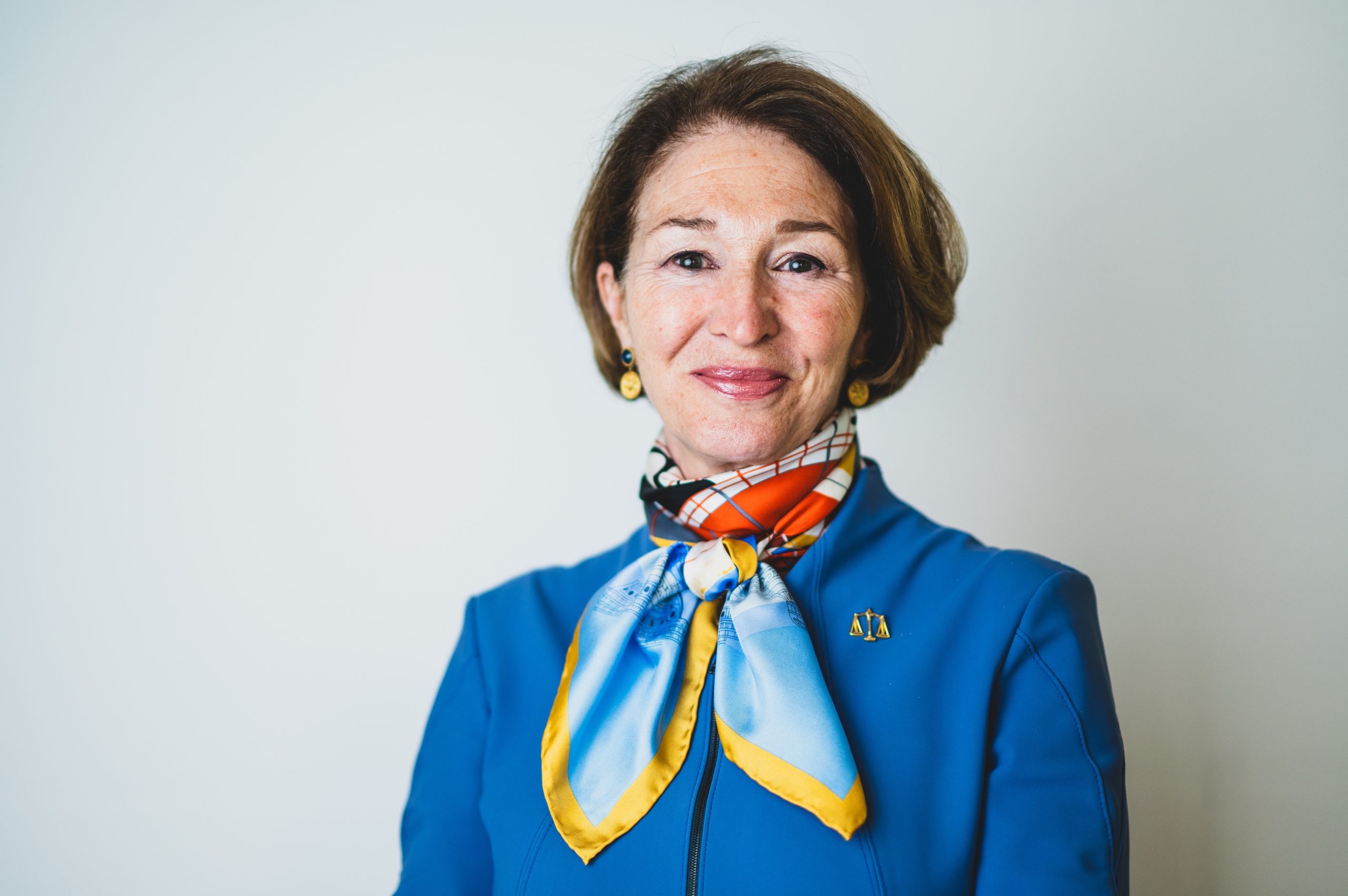 A smiling woman with short brown hair wearing a blue jacket, gold earrings, and a colorful scarf stands in front of a plain white background.
