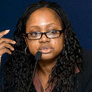A woman with braided hair and glasses speaks into a microphone. She is wearing a dark blazer and hoop earrings, and sits in front of a dark blue background. One hand is raised near her face.