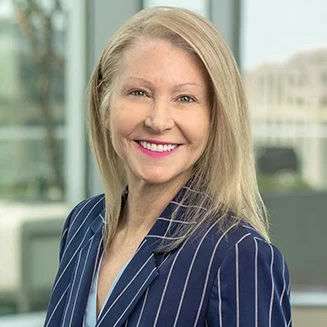 Smiling woman with long blonde hair, wearing a blue pinstripe blazer, standing indoors in front of large windows with a blurred office background.