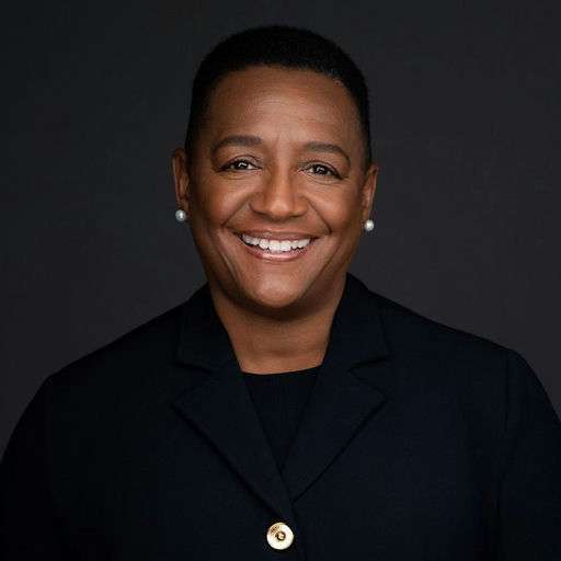 A person with short hair and pearl earrings wears a dark suit jacket over a top, smiling against a plain dark background.