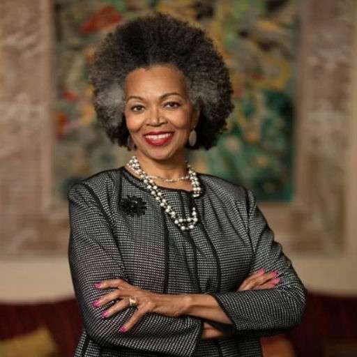 A confident older woman with gray afro hair, wearing a black dress, pearls, and a brooch, stands with arms crossed and smiling in a warmly lit room with colorful artwork in the background.
