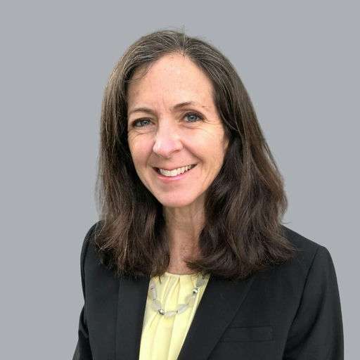 A woman with long brown hair wearing a black blazer and a light yellow blouse smiles at the camera against a plain gray background.