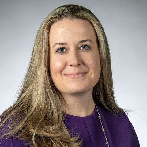 A woman with long blonde hair wearing a purple top and a necklace, smiling at the camera against a plain gray background.