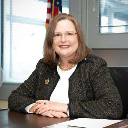 A woman with shoulder-length brown hair, glasses, and a gold necklace is sitting at a desk, smiling, with hands folded. She is wearing a dark blazer over a white top. An American flag is visible in the background.