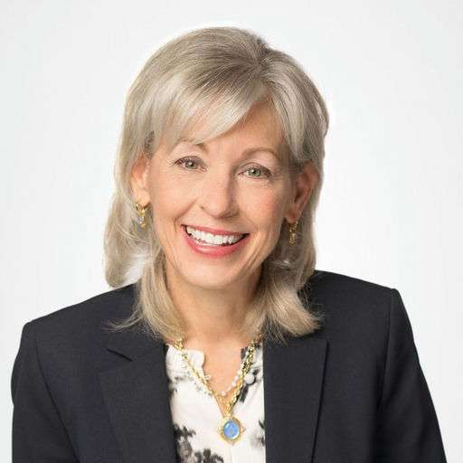 A woman with shoulder-length gray hair smiles, wearing a dark blazer, a light-patterned blouse, and gold jewelry, including earrings and a necklace with a blue pendant, against a plain light background.