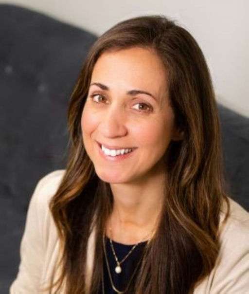 A woman with long brown hair, wearing a beige blazer over a dark top and layered necklaces, smiles while seated on a dark gray couch.