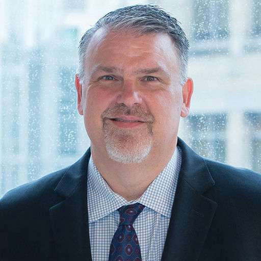 A man with short gray hair and a goatee, wearing a dark suit, white shirt, and patterned tie, smiles in front of a window with a blurred cityscape in the background.