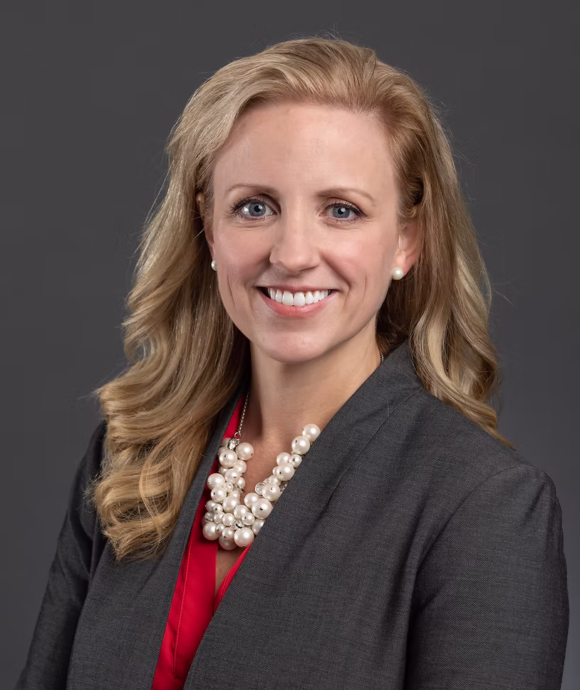 A woman with long, wavy blonde hair wearing a dark gray blazer, red blouse, and a large pearl necklace, smiling in front of a plain gray background.