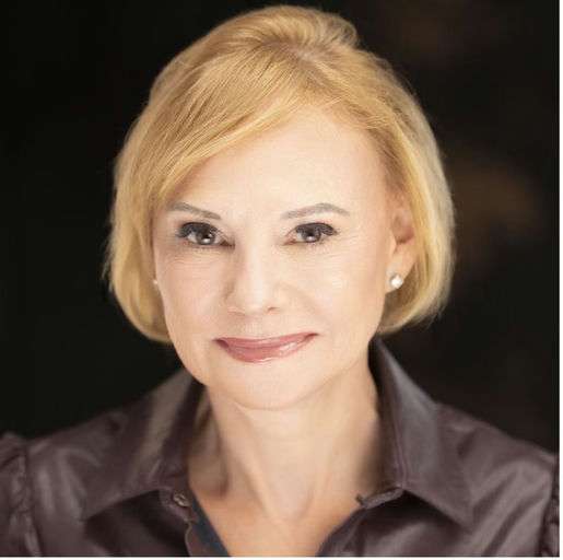 A woman with short blonde hair, light skin, and wearing a dark collared shirt smiles gently at the camera against a dark, blurred background.