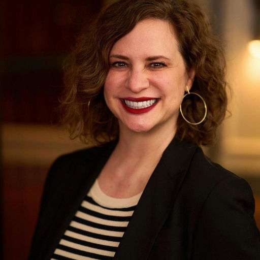 A woman with wavy brown hair, wearing large hoop earrings, a black blazer, and a black-and-white striped top, smiles at the camera against a softly blurred background.