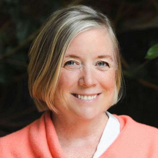 A woman with short, light blonde hair and fair skin smiles at the camera. She is wearing a coral-colored top, and there are green, blurred plants in the background.
