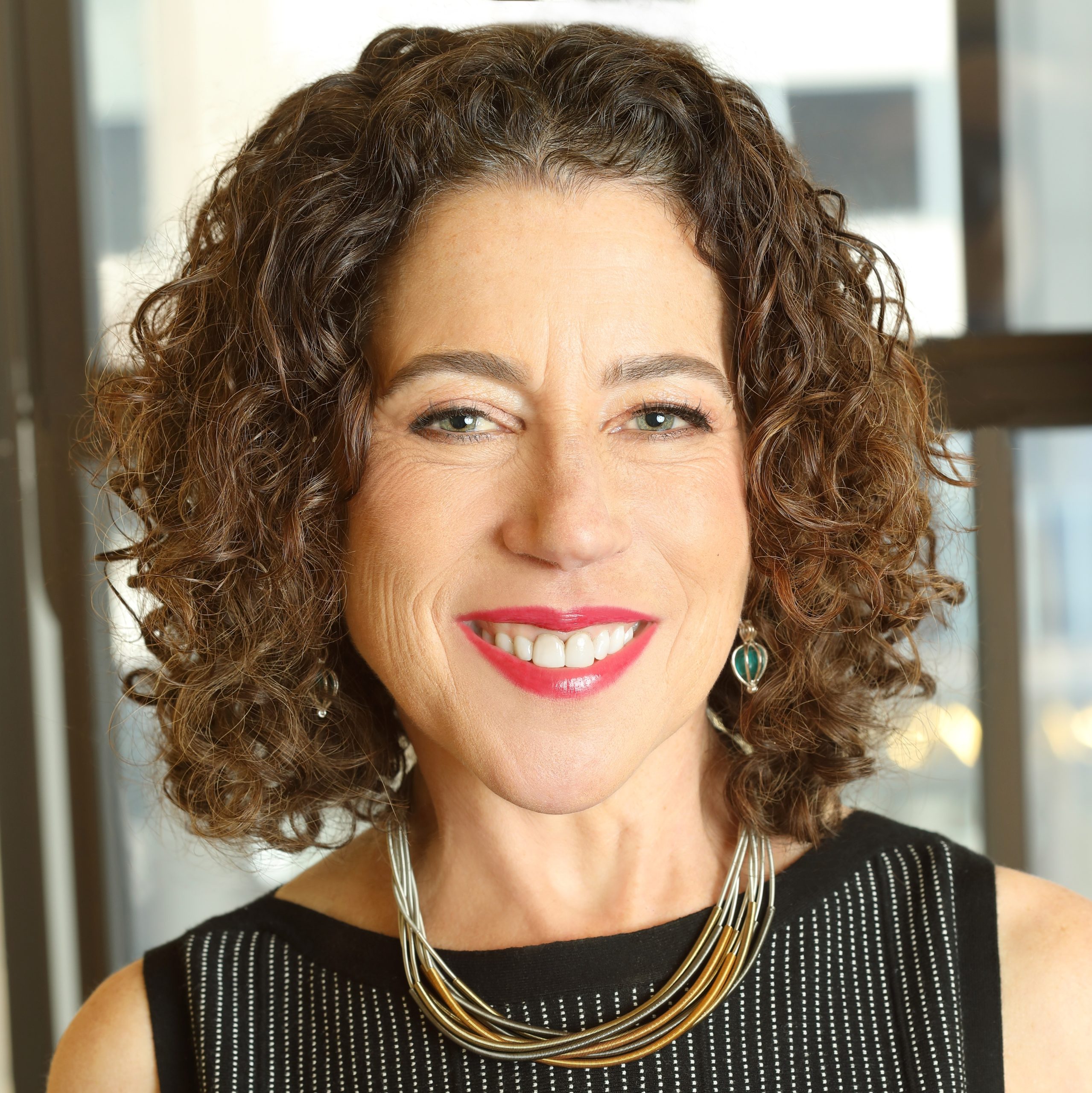 A woman with curly brown hair, wearing a sleeveless black dress with white stripes, silver hoop earrings, and a layered silver necklace, smiling in front of a window with blurred background.
