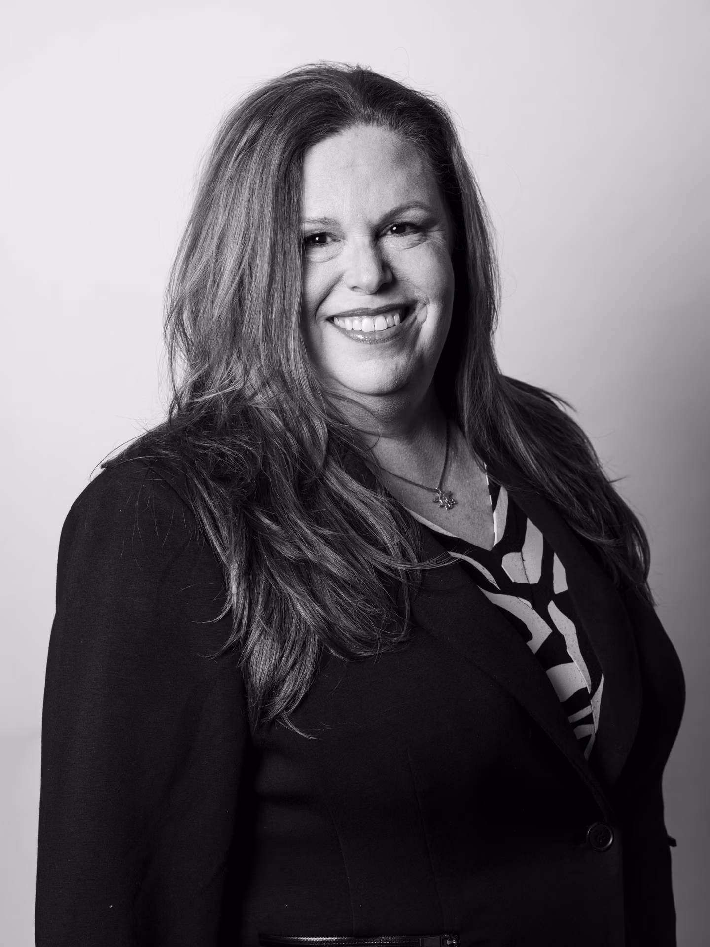 A woman with long hair smiles while wearing a dark blazer over a patterned top. The photo is black and white with a plain background.