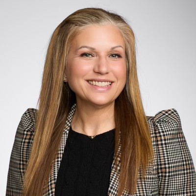 A woman with long straight blonde hair wearing a plaid blazer over a black top, smiling at the camera against a plain light gray background.